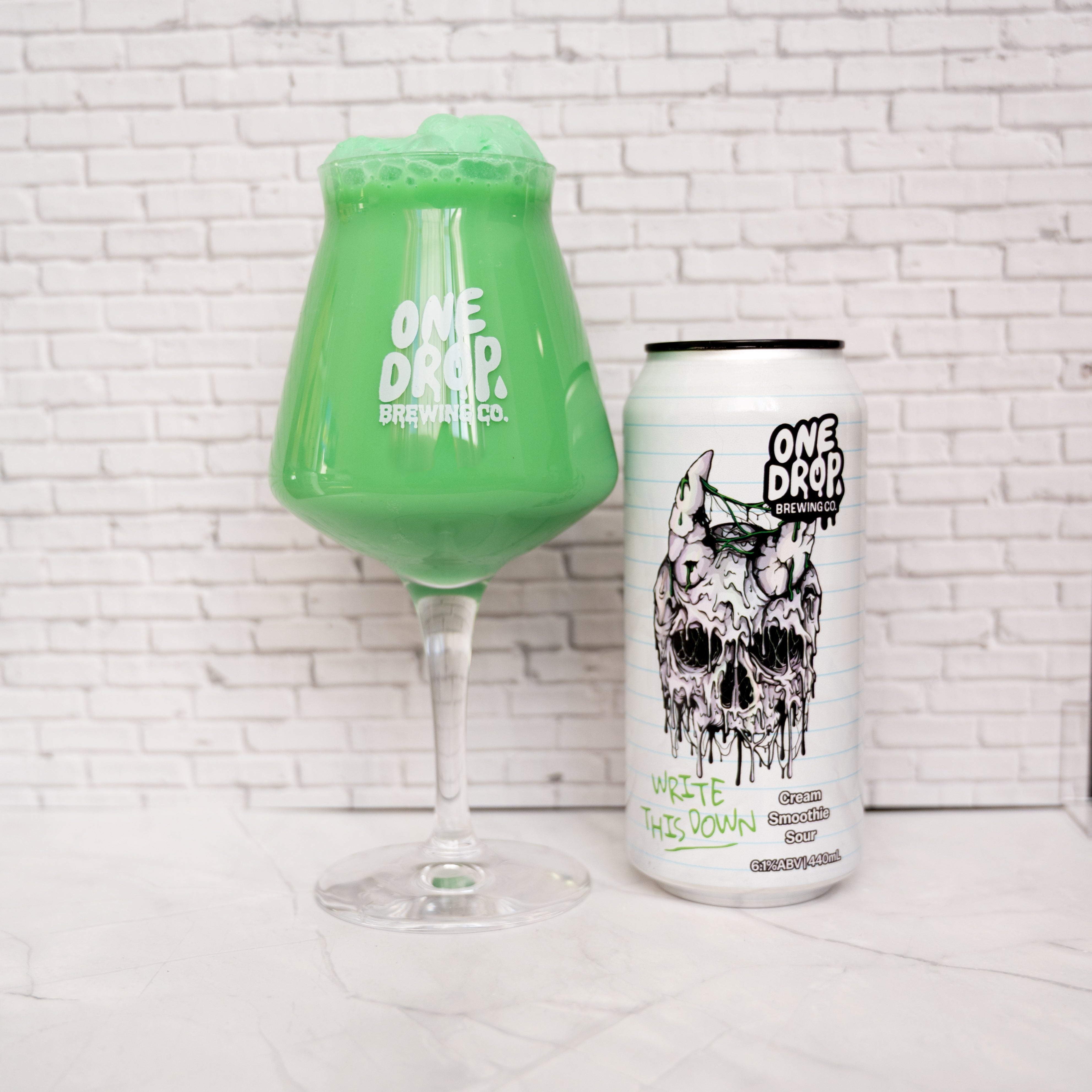 A Glass full of green write this down beer for  of One Drop Brewing Co beside his can.  set against a white brick wall background.